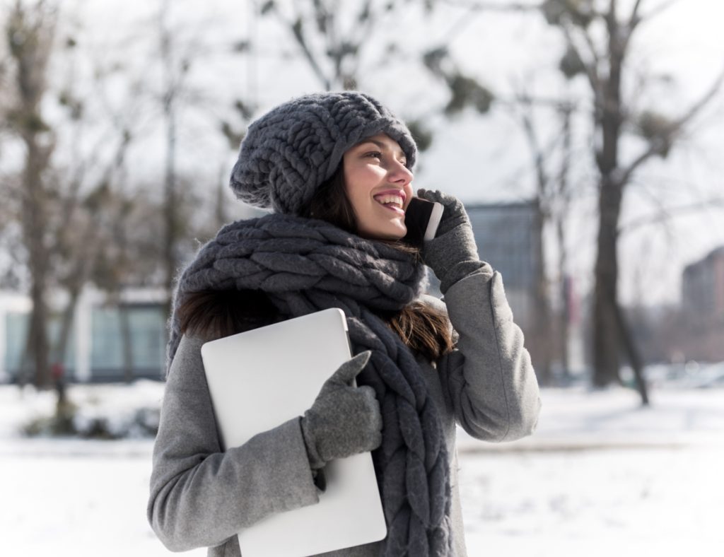 Woman with laptop under arm and speaking on phone outside in winter.